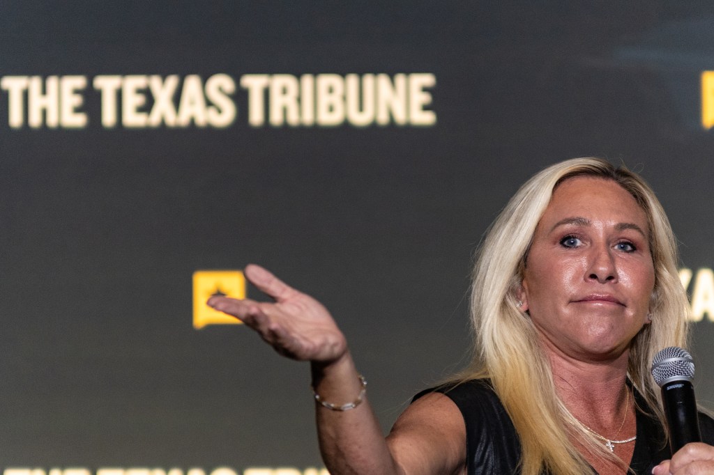 A woman speaking into a microphone with a serious expression, gesturing with her hand, in front of a screen displaying 'The Texas Tribune.'