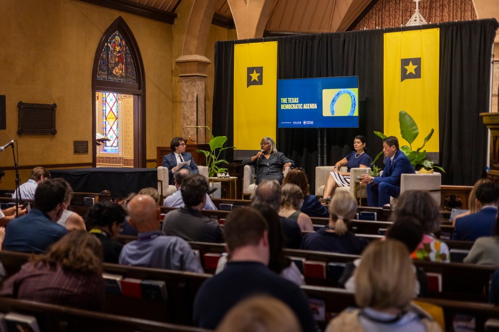 A panel discussion featuring four speakers on stage discussing the Texas Democratic Agenda, with an audience seated in a historic venue adorned with decorative banners.