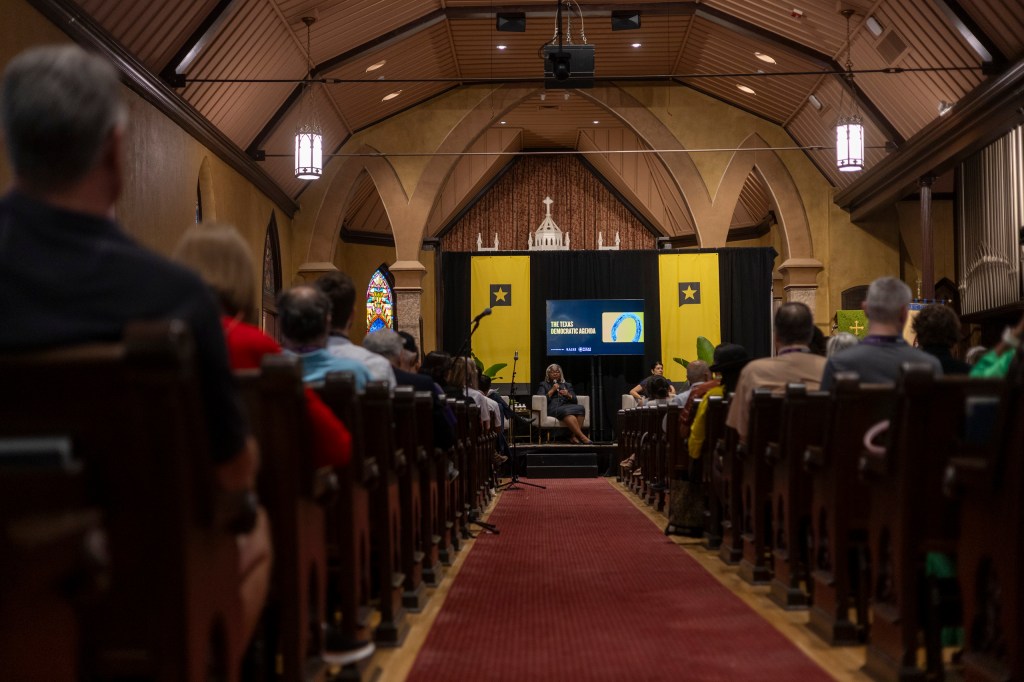 A diverse audience seated in a church interior, facing a stage with a presentation screen and speakers during an event.