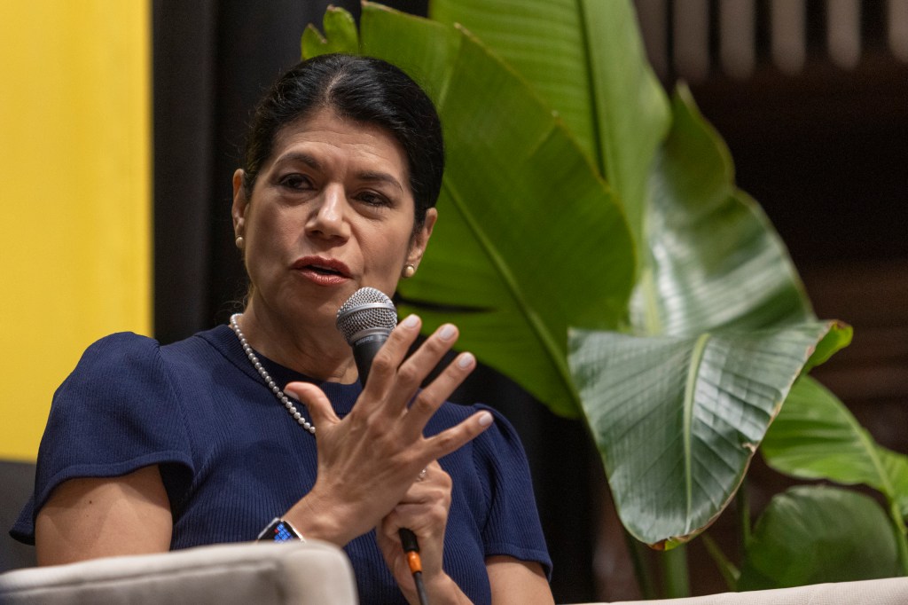 A woman in a navy blue shirt speaks into a microphone, gesturing with her hands, in front of a backdrop featuring large green leaves.
