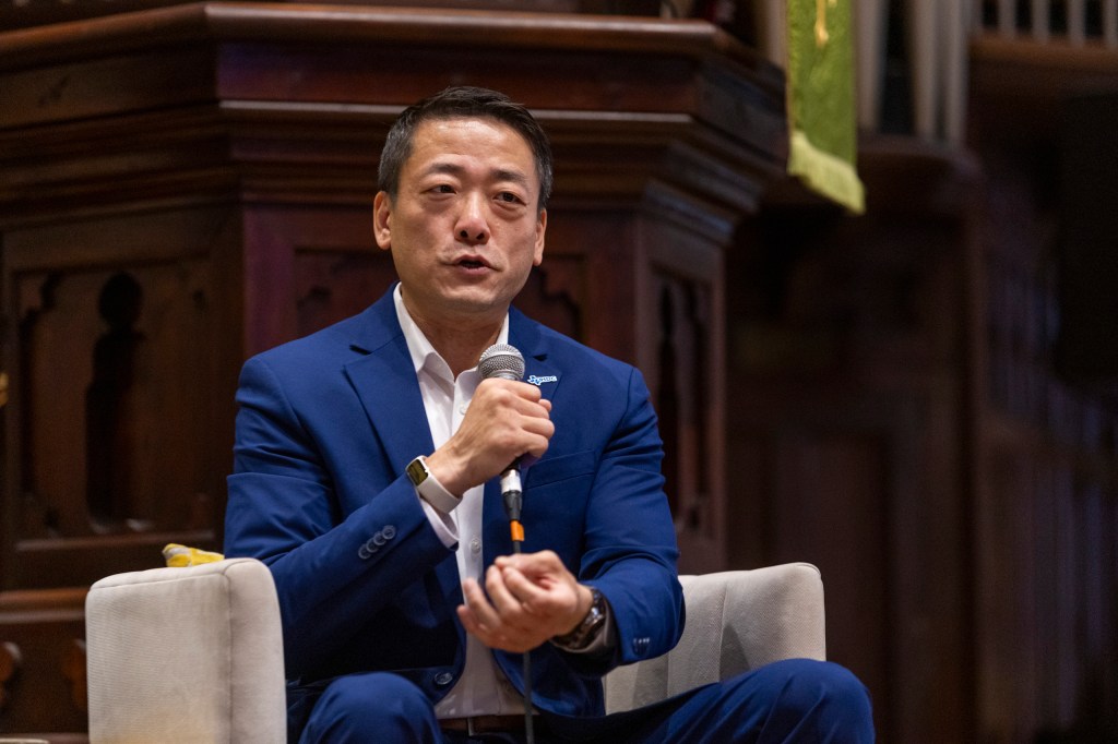 A man in a blue suit speaking into a microphone while seated on a stage with wooden paneling in the background.