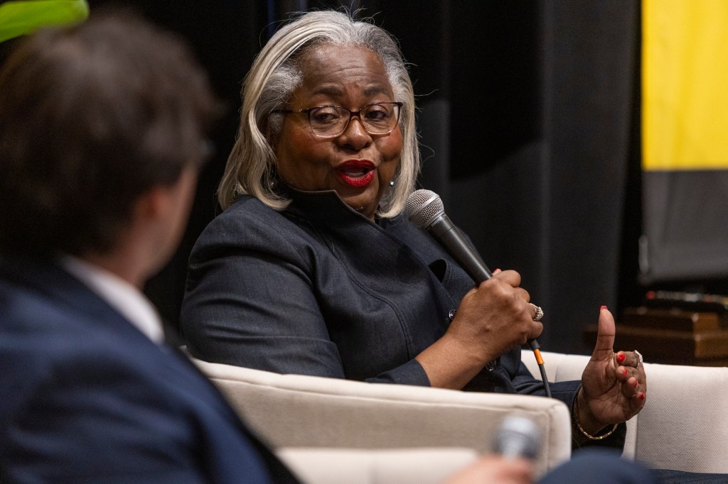 A woman with gray hair speaking into a microphone during a panel discussion, seated comfortably in a chair, while a man in business attire listens attentively.