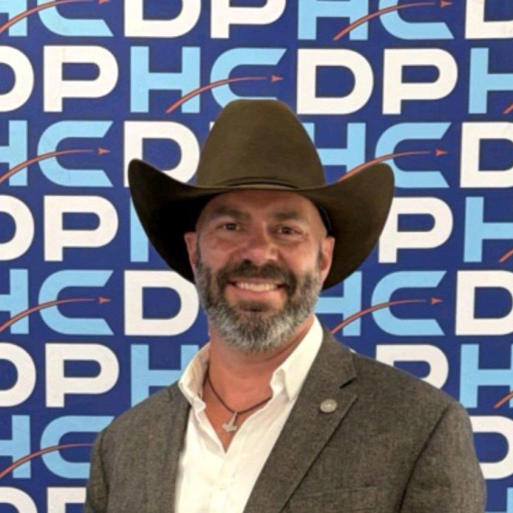 Man with a beard wearing a brown cowboy hat and a blazer, smiling in front of a blue and white patterned background.