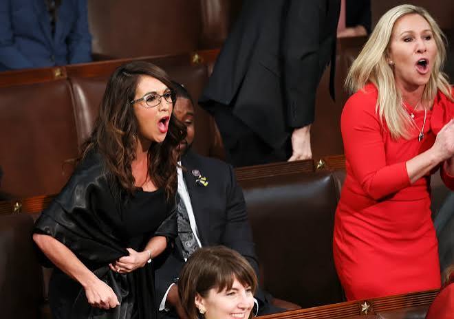Two women expressing strong emotions while standing in a legislative assembly, with one wearing a black dress and the other in a red dress.