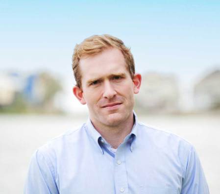 A man with short light brown hair, wearing a light blue button-up shirt, stands outdoors with a soft-focus beach background.
