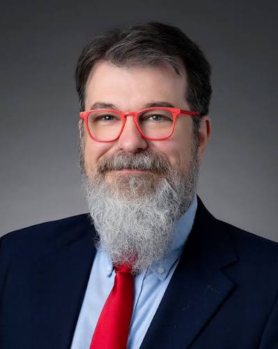 A middle-aged man with a beard and glasses, wearing a suit and a red tie, poses for a professional portrait against a gray background.