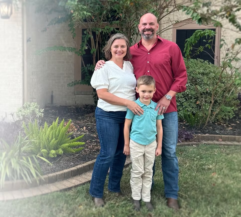 A family portrait featuring two adults, a man and a woman, standing with a young boy in front of a house and garden. The man is wearing a red shirt and the woman is in a white top with jeans, while the boy is dressed in a light blue shirt and beige pants.